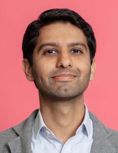 A person with short dark hair, wearing a gray blazer and light blue collared shirt, is looking at the camera against a pink background.